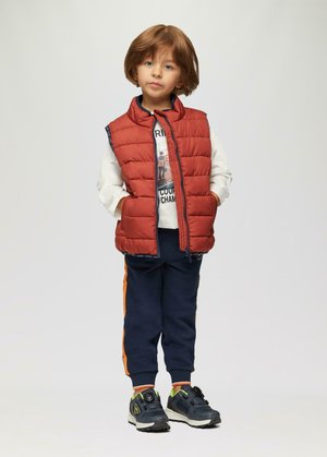 Rote, bauschige Weste über einem weißen Hemd, marineblaue Sweatpants mit orangen Seitenstreifen und schwarze Sportschuhe mit neonfarbenen Akzenten. Kind steht mit den Händen in den Taschen.