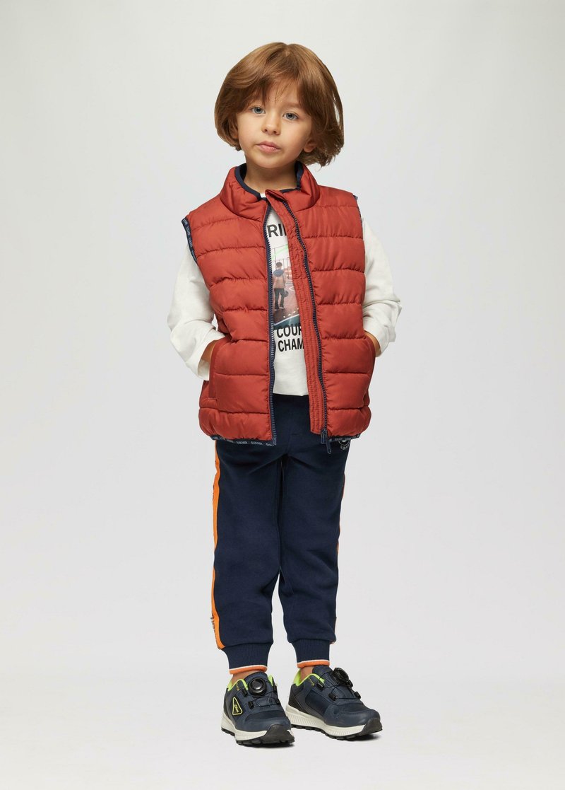 Rote, bauschige Weste über einem weißen Hemd, marineblaue Sweatpants mit orangen Seitenstreifen und schwarze Sportschuhe mit neonfarbenen Akzenten. Kind steht mit den Händen in den Taschen.