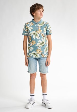 Kurzärmliges, türkisfarbenes Poloshirt mit weißem und orangefarbenem Blumenmuster, kombiniert mit hellen Denim-Shorts und weißen Sneakers mit schwarzen Akzenten.
