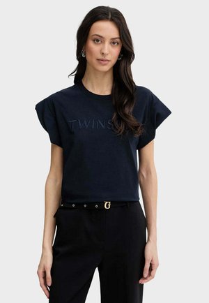 Donna con lunghi capelli scuri che indossa un top nero senza maniche con la scritta "TWINS" e pantaloni neri con cintura nera con fibbia dorata a forma di "G".