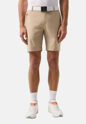 Mann trägt beige Shorts mit Reißverschlusstasche an der Seite, weißen Gürtel, weiße Socken und weiße Turnschuhe mit orangefarbenen Schnürsenkeln und steht vor einem einfarbigen Hintergrund.