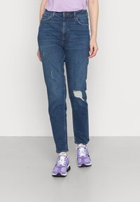 Jeans azuis de cintura alta com um corte ligeiramente ajustado, apresentando detalhes desgastados e um design clássico de cinco bolsos, combinados com ténis roxos.