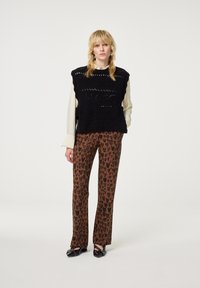 Blonde vrouw staat en draagt een zwart mouwloos gebreid vest, een crèmekleurig shirt met lange mouwen, bruine broek met luipaardprint en zwarte puntige schoenen.