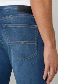 Dark blue denim jeans with a back pocket featuring red, white, and blue logo detail and contrasting stitching, shown from the side.
