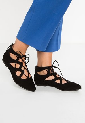 Stopy w czarnych zamszowych płaskich butach na sznurówki z wycięciami, w połączeniu z niebieskimi, krótkimi szerokimi spodniami na białej podłodze.