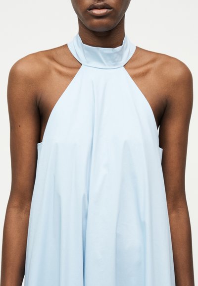 Femme portant une robe bleu clair sans manches à col halter haut, montrée des épaules au milieu du torse sur un fond blanc.