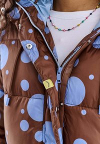 Kurtka puchowa z brązowej tkaniny z dużymi niebieskimi kropkami. Posiada jasno niebieski kaptur, zapięcie na suwak oraz żółtą metkę z wzorem oka.