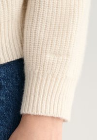 Jersey de punto color crema con puños acanalados, que presenta un sutil logo bordado en la manga, combinado con vaqueros de mezclilla azul oscuro.