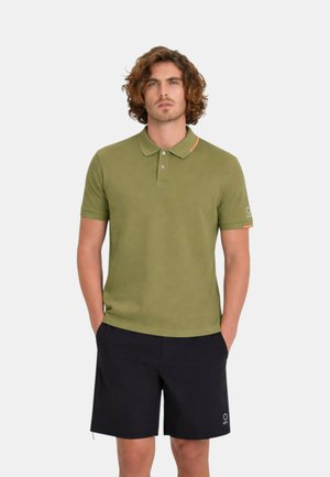 Uomo con capelli ricci che indossa una polo verde oliva e pantaloncini neri, in piedi con le mani in tasca davanti a uno sfondo semplice.