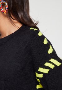Svart stickad tröja med texturerad tyg, med klara gula ränder på ärmarna. Nära bild ger detaljer om halslinningen.