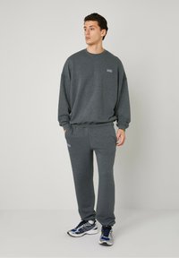 Sweatshirt gris et pantalon de jogging assorti en tissu texturé, avec un petit logo sur la poitrine. Porté avec des baskets bleues et blanches.