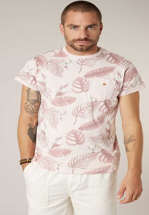 Homme aux cheveux courts et barbe portant un t-shirt blanc à imprimé feuilles tropicales roses et blanches, et un pantalon blanc, debout les mains détendues.