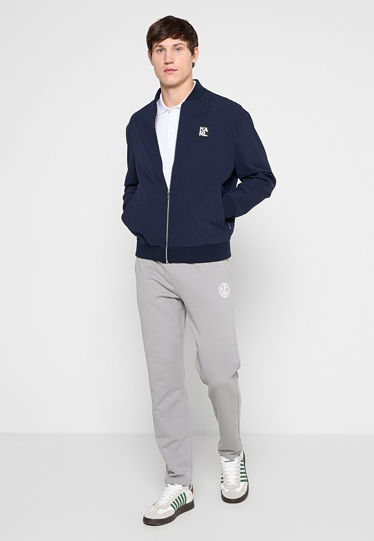 Joven de pie con las manos en los bolsillos, lleva una chaqueta azul marino con cremallera, una camiseta polo blanca, pantalones de chándal grises y zapatillas blancas con rayas verdes.