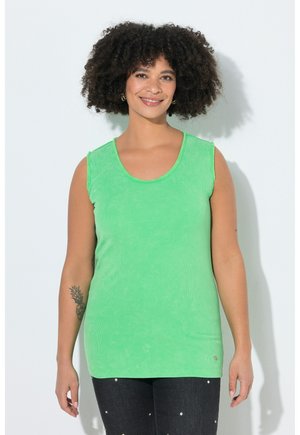 Vrouw met krullend haar die glimlacht, draagt een mouwloze groene top en zwarte broek met kleine witte stippen, staand tegen een effen achtergrond.