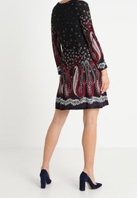 Vestido negro con mangas largas, que presenta patrones florales y de paisley en rojo y blanco en el dobladillo, combinado con tacones altos azul marino.