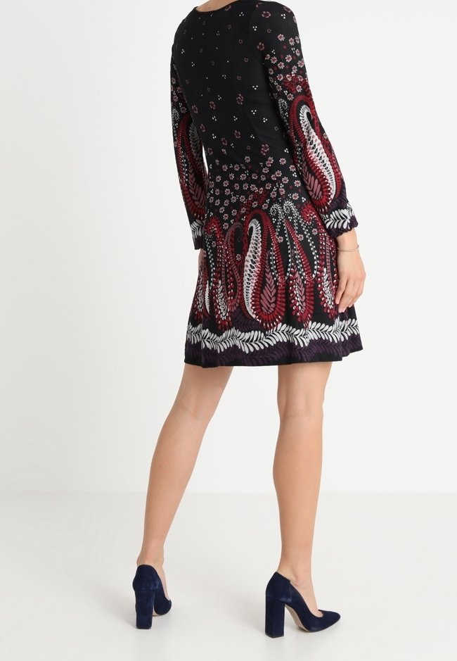 Vestido negro con mangas largas, que presenta patrones florales y de paisley en rojo y blanco en el dobladillo, combinado con tacones altos azul marino.