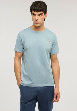 Giovane uomo con capelli ricci scuri che indossa una maglietta a maniche corte azzurro chiaro e pantaloni blu scuro, in piedi con le mani in tasca.