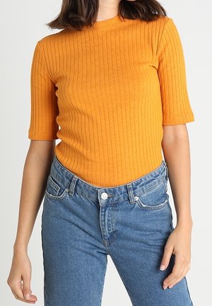 Kobieta ubrana w pomarańczowy, prążkowany T-shirt z krótkim rękawem, włożony do wysokich niebieskich dżinsów, stoi z jedną ręką swobodnie opuszczoną wzdłuż ciała.