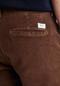 Ruskeat corduroy-housut sisältävät rakenteellisen pinnan, takataskun ja valkoisen merkkilapun, jossa lukee "Pepe Jeans London".
