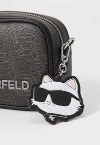 Črna teksturirana torba z sivo embosirano zasnovo. Vsebuje bel mačji obesek s črnimi akcenti in sončnimi očali, pritrjen z varnostnim sponko.