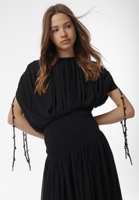 Schwarzes Kleid mit puffierten kurzen Ärmeln, einem gesmokten Oberteil und einem gerafften Rock. Verfügt über dekorative schwarze und goldene Akzente an den Zugbändern.