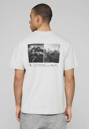 Man die een lichtgrijs T-shirt draagt met twee zwart-wit abstracte foto's en kleine tekst op de achterkant, staand tegen een effen achtergrond.