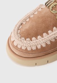 Scarpa in suede marrone con cuciture intrecciate beige, suola piatta di colore chiaro e dettaglio dorato sul lato.