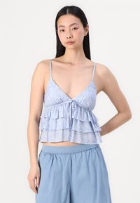 Svetlo modrý top s viacerými vrstvami a tenkými ramienkami, so zaviazaním vpredu a paisley vzorom na mäkkom materiáli. Spárované s džínsovými nohavicami.