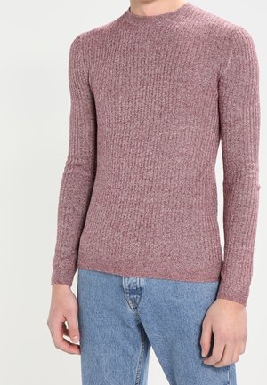 Uomo che indossa un maglione aderente a maniche lunghe in maglia costina color malva abbinato a jeans di denim chiaro su uno sfondo semplice.