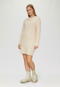 Strickkleid in hellem Beige mit einem strukturierten Muster aus ovalen Formen, langen Ärmeln und einem gerippten Saum. Kombiniert mit beigen Knöchelstiefeln.