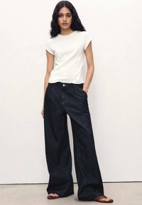 T-shirt blanc à manches courtes associé à un pantalon en denim large bleu foncé, doté d'une poche avant et de détails de couture visibles.