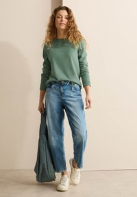 Grüner Pullover mit gerippten Bündchen, hellblaue Jeans, weiße Sneaker und eine weiche grüne Tasche. Lässiger, entspannter Schnitt.