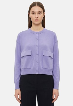 Donna che indossa un cardigan lilla chiaro con bottoni e due tasche frontali con patta, e pantaloni neri, in piedi contro uno sfondo semplice.