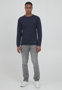 Pulover texturat de culoare bleumarin cu mânecă lungă și guler rotund, asortat cu blugi gri deschis și adidași albi, stând pe un fundal simplu.