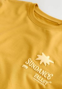 T-shirt jaune à col rond avec texte blanc en relief « Sundance Valley 1999 California Sounds » et un motif en forme d'étoile au-dessus du texte.