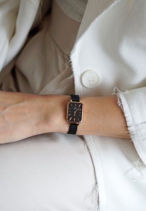 Montre rectangulaire noire avec des accents en or rose et un bracelet en tissu texturé, portée au poignet. Veste blanche avec des bords effilochés partiellement visible.