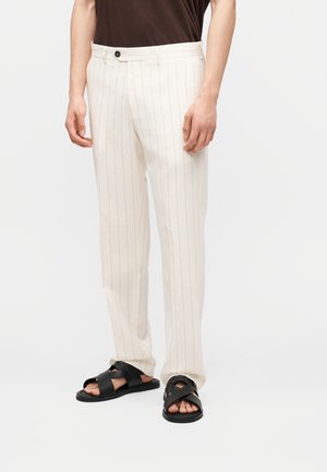 Man wearing cream-colored pinstripe trousers and black leather cross-strap sandals, standing against a plain background.