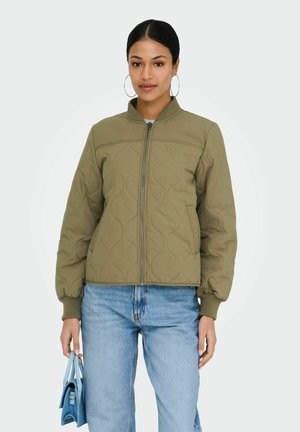 Femme portant une veste bomber matelassée olive, jean bleu clair, grandes boucles d'oreilles anneaux, tenant un petit sac à main bleu à ses côtés.