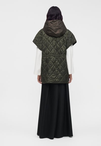 Personne portant une veste matelassée sans manches à capuche vert foncé par-dessus des manches longues blanches et un pantalon noir à jambes larges, vue de dos.