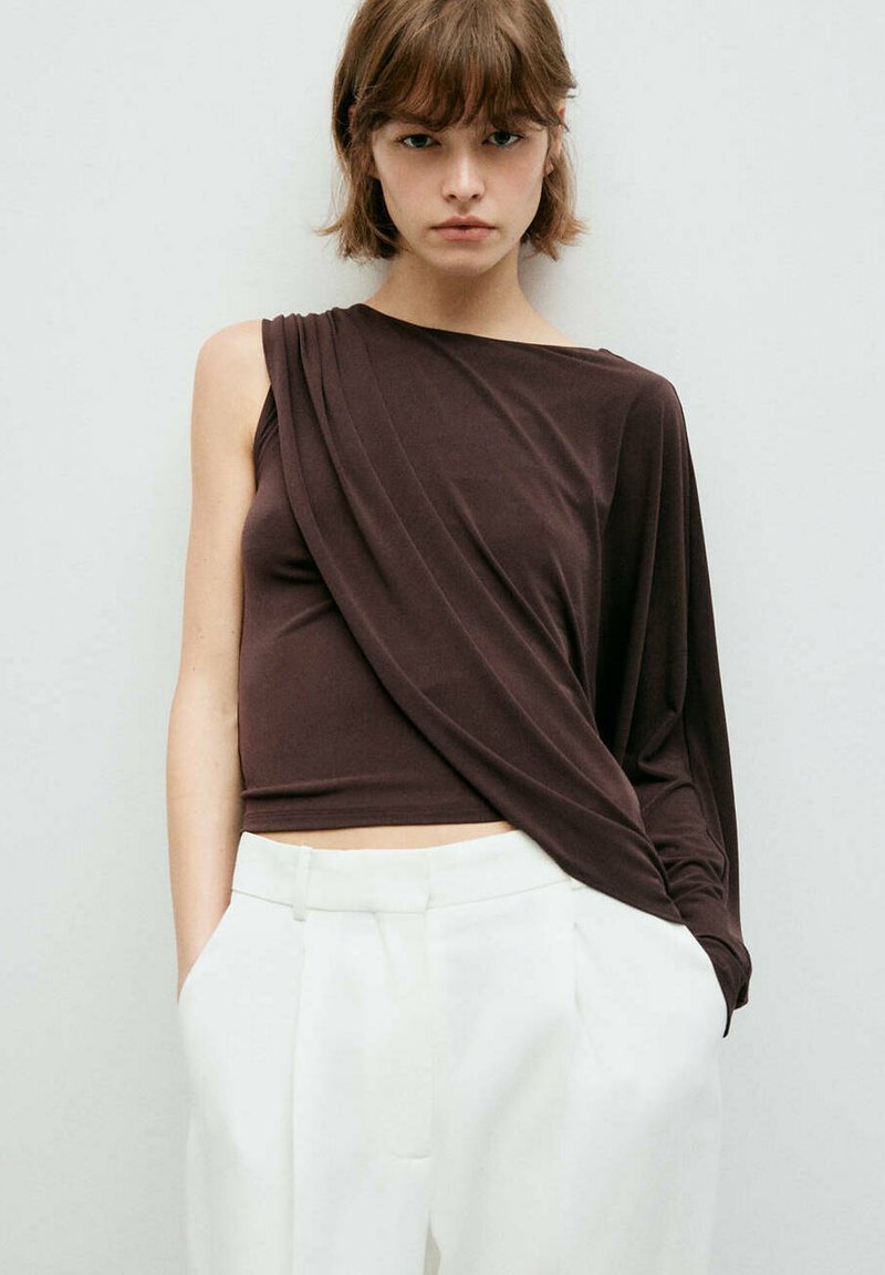 Jeune femme aux cheveux courts portant un haut asymétrique à manches longues marron foncé et un pantalon blanc taille haute, debout contre un mur uni.