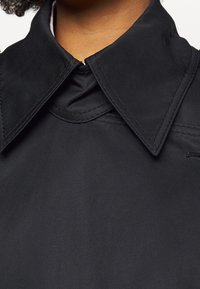 Czarny, strukturalny kołnierz płaszcza, wykonany z gładkiego, matowego materiału, z widocznym przeszyciem i subtelną metką marki z boku.