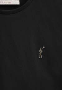 Svart t-shirt med rund halsringning, tillverkad av mjukt bomullstyg. Har ett emblem av en golfsäljare i guld strass på vänster bröst.