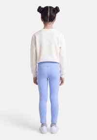 Παιδί με δύο κότσους, φορώντας λευκό φούτερ, ανοιχτό μπλε κολάν και λευκά αθλητικά, στέκεται με την πλάτη γυρισμένη σε απλό φόντο.