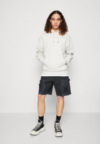 Felpa grigia chiara con cappuccio e lacci, tasca frontale e logo ricamato, abbinata a pantaloni cargo scuri e sneaker alte nere.