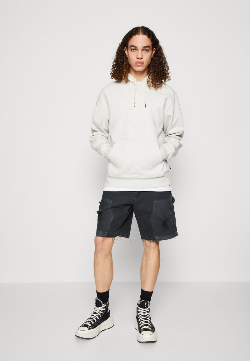 Felpa grigia chiara con cappuccio e lacci, tasca frontale e logo ricamato, abbinata a pantaloni cargo scuri e sneaker alte nere.