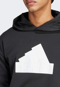 Sudadera negra con un logo de Adidas en blanco texturizado, con capucha ajustable y una tela suave y lisa. Sin patrones ni acentos visibles.
