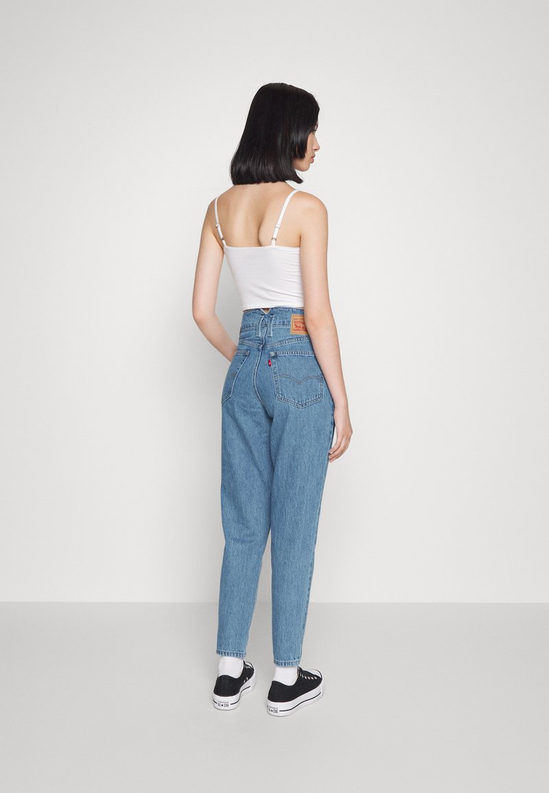 Jeans de mezclilla azul de talle alto con un corte relajado, que cuentan con una etiqueta de cuero en la parte trasera, bolsillos laterales y dobladillos. Combinados con zapatillas negras.