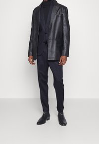 Černý texturovaný kožený blazer s klopovými límci, v kombinaci s tmavým oblekem a přiléhavými kalhotami, doplněný černými kotníkovými botami.