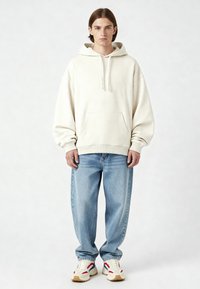 Felpa oversize color crema con tasca frontale, abbinata a jeans in denim blu chiaro e sneakers bianche con dettagli rossi e blu.