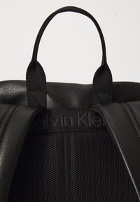 Sac à dos en cuir noir avec une surface texturée, des bretelles rembourrées et une poignée supérieure, doté d'une marque discrète en noir à l'intérieur.